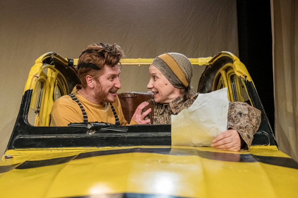 Zwei Schauspieler*innen in einem gelb-schwarzen Auto, die sich in einem intensiven Gespräch befinden.