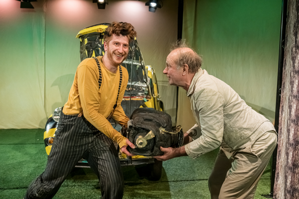 Zwei Schauspieler halten gemeinsam einen Motor mit einem gelb-schwarzen Auto im Hintergrund.