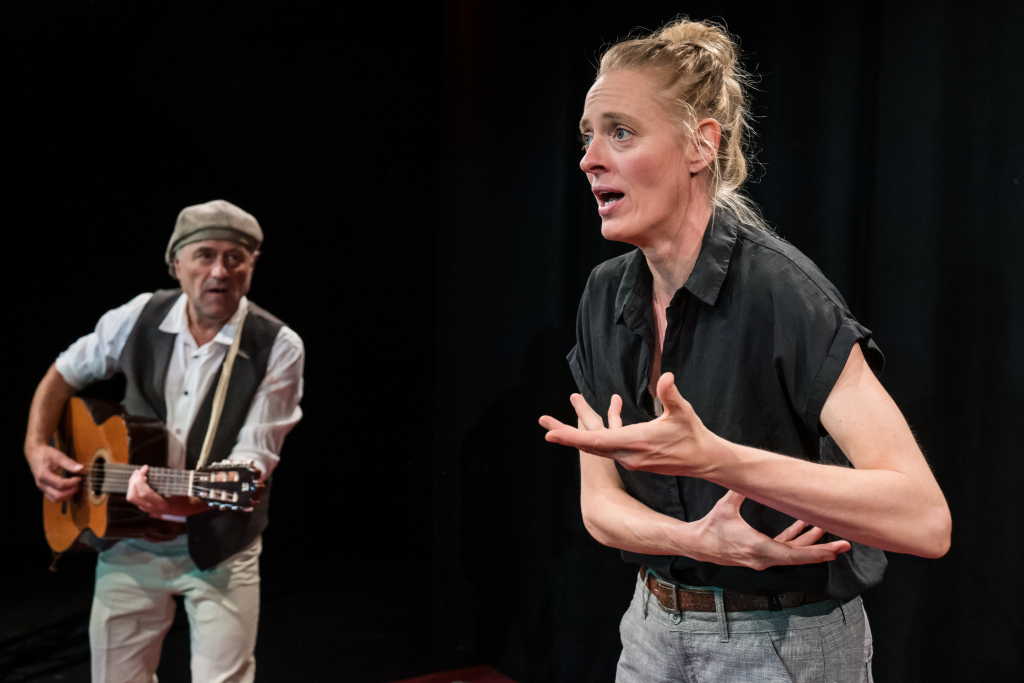 Frau mit hochgestecktem Haar und schwarzem Hemd gestikuliert mit beiden Händen, im Hintergrund Mann mit weißem Hemd und Weste spielt Gitarre.