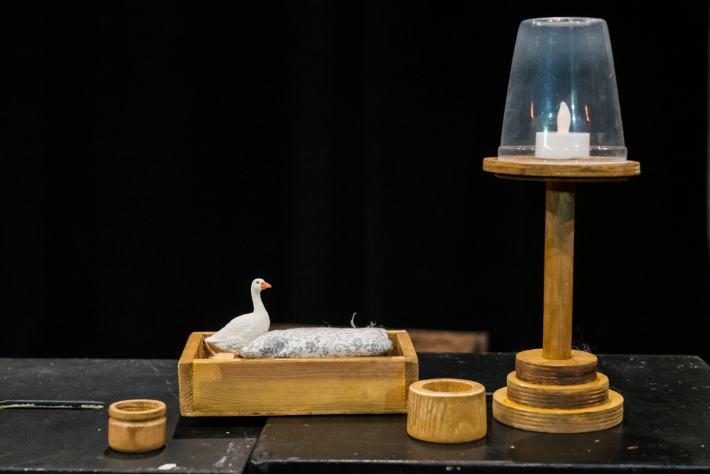 Holztablett mit einer weißen Gansfigur auf einem grauen Kissen, daneben zwei kleine Holzgefäße und ein Holzständer mit brennender Kerze unter Glas.