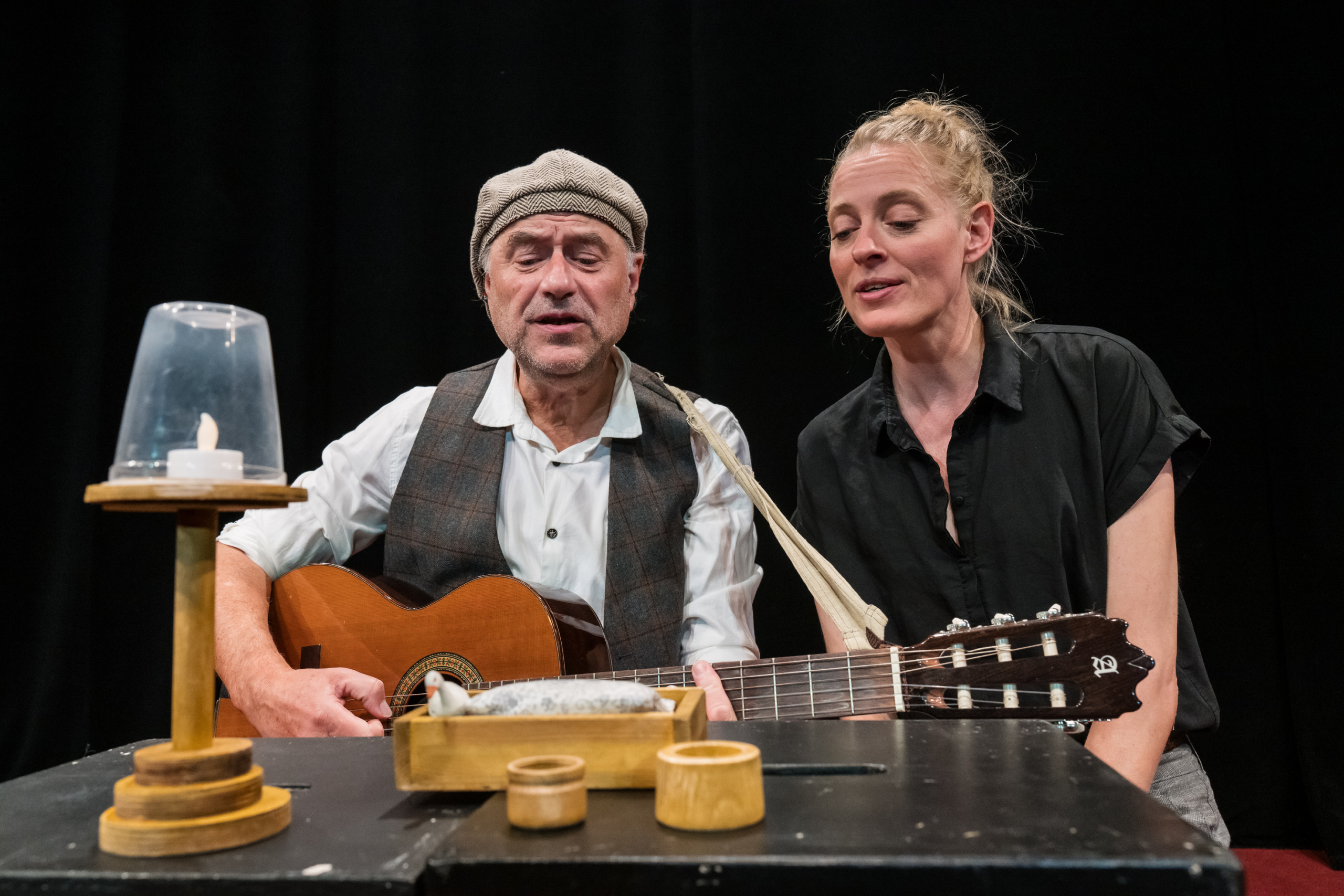 Zwei Personen sitzen an einem Tisch, eine spielt Gitarre, auf dem Tisch stehen ein Kerzenständer mit Glas und mehrere kleine Holzgefäße.