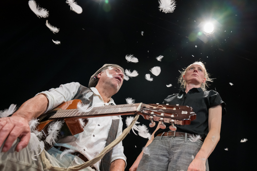 Zwei Personen, eine mit Gitarre, umgeben von schwebenden weißen Federn vor dunklem Hintergrund.