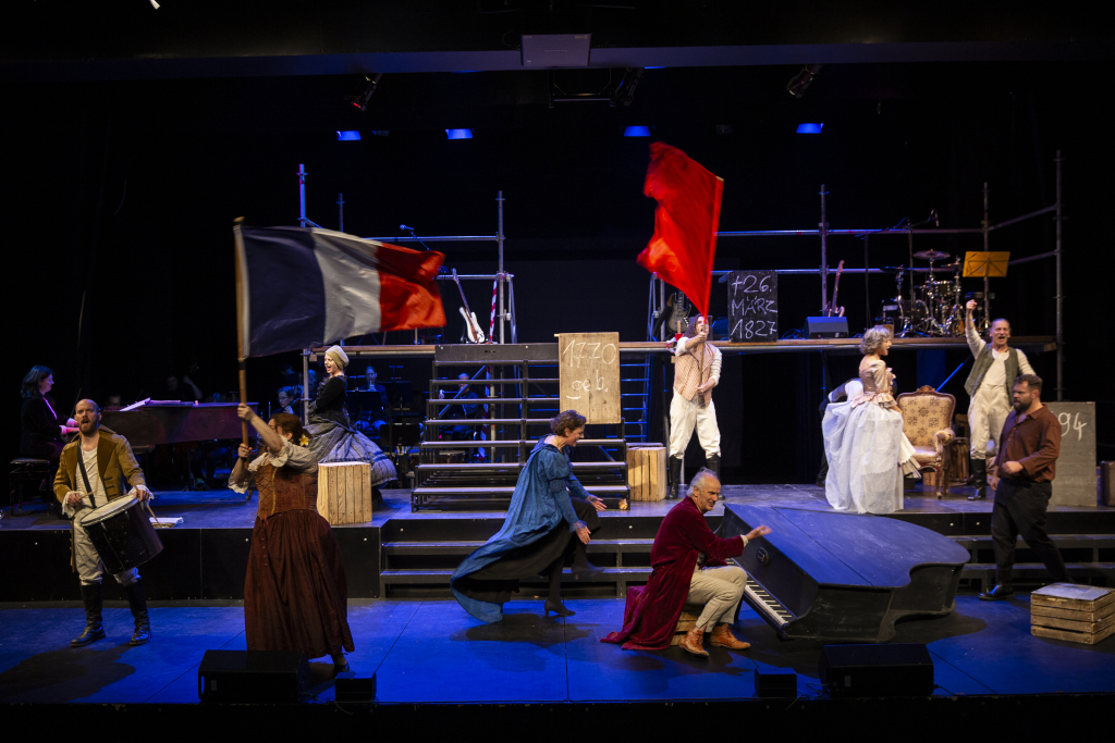 Bühnenbild mit mehreren Schauspielern in historischen Kostümen, zwei halten große Fahnen, im Hintergrund Schlagzeug und Treppe.