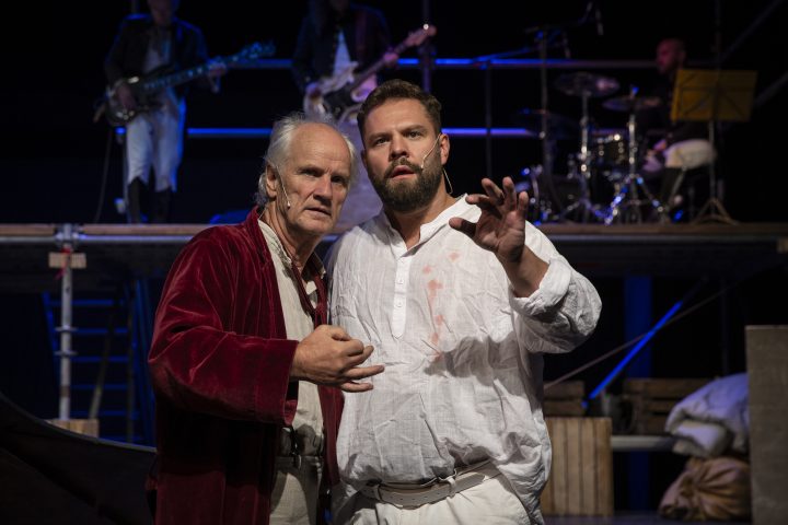 Zwei Männer in historischen Kostümen, einer in rotem Samtjackett, der andere in weißem Hemd, vor einer Bühne mit Musikern.