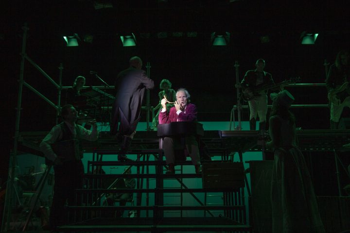 Bühnenbild mit mehreren Personen, darunter eine sitzende Person in rotem Oberteil, umgeben von weiteren Personen in dunkler Kleidung und Musikinstrumenten.