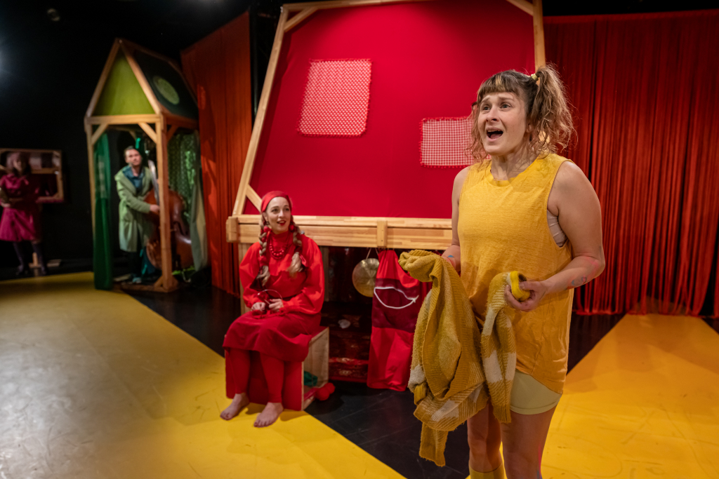 Schauspielerin in gelbem ärmellosem Oberteil und Shorts hält gelbe Jacke, im Hintergrund zwei Personen in roter und grüner Kleidung vor rotem Bühnenbild.