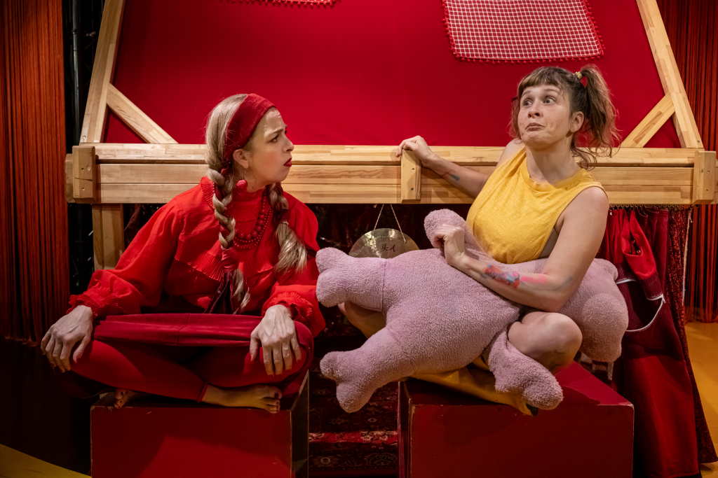 Zwei sitzende Schauspielerinnen vor rotem Hintergrund, eine trägt rotes Kleid und Perlenkette, die andere gelbes ärmelloses Oberteil und hält ein großes rosa Stofftier.