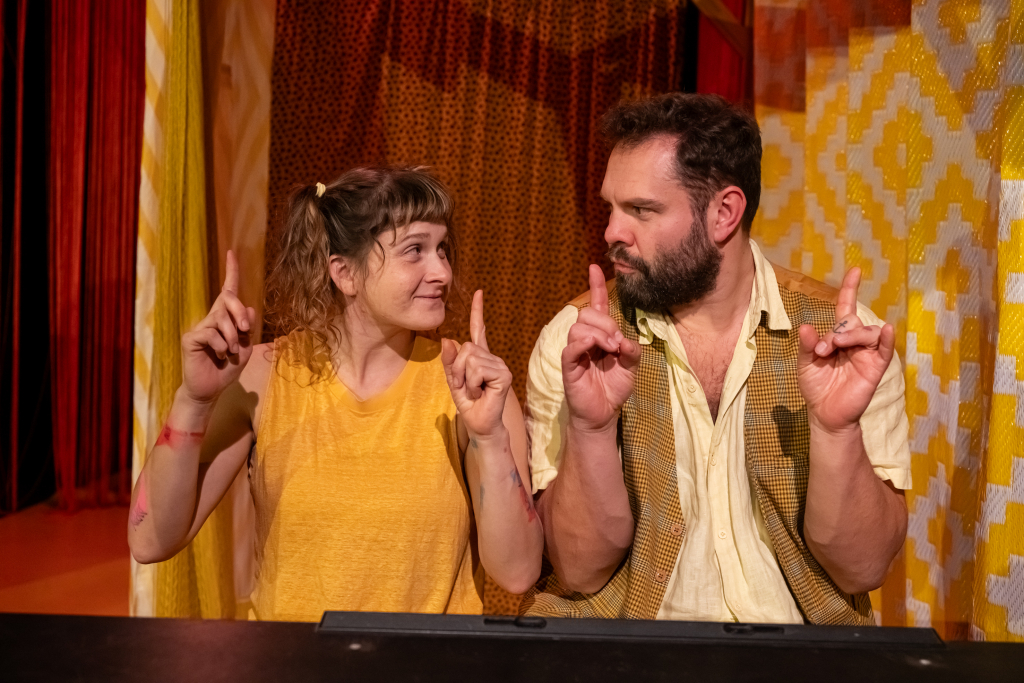 Zwei Personen sitzen nebeneinander, heben jeweils beide Zeigefinger und schauen sich an, im Hintergrund gelb-rote Vorhänge mit Mustern.