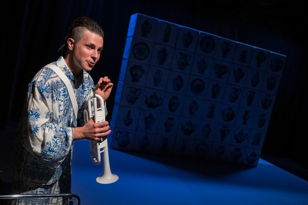 Mann in weiß-blauen Anzug mit Trompete auf der Bühne vor einem dekorativen Wandbild.