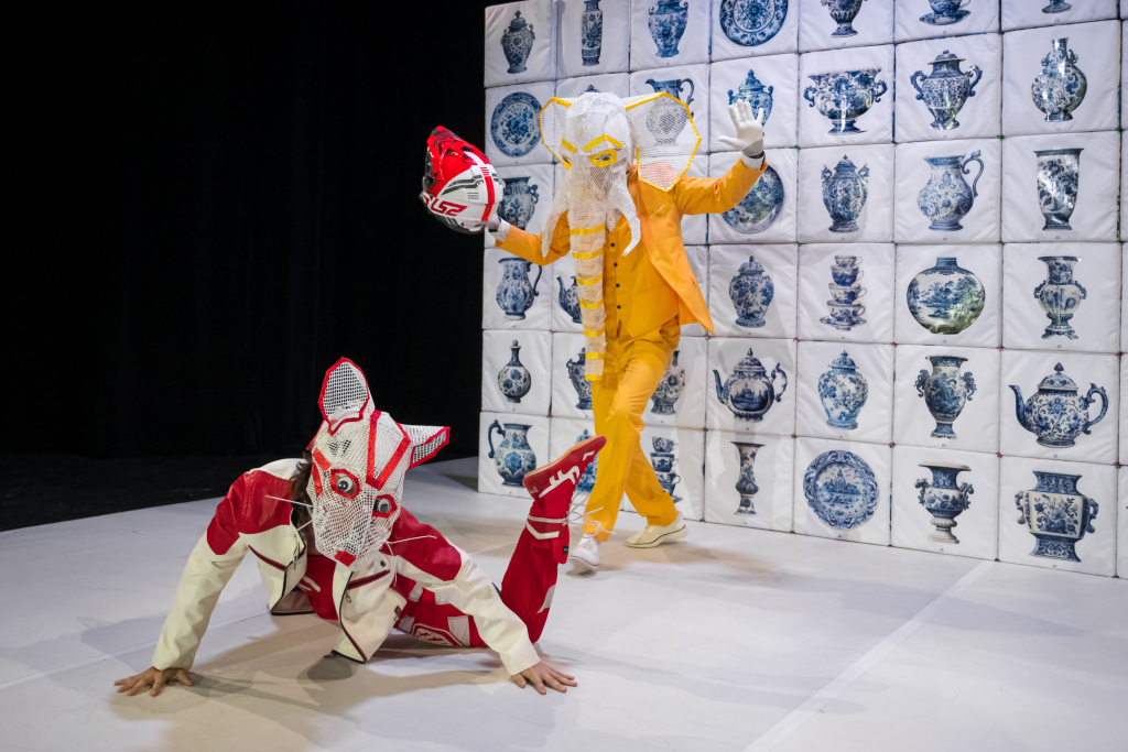 Zwei Schauspieler*innen mit Masken, einmal in rot und einmal in gelb, auf einer Bühne vor einer Wand mit balu-weißer Bemalung.