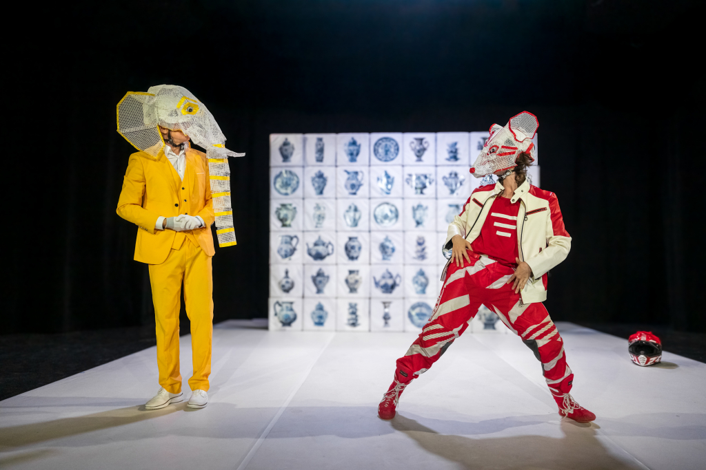 Zwei Schauspieler*innen in kreativen Kostümen: gelber Anzug mit Elefantenmaske und rot-weißes Outfit mit Mäusemaske auf einer Bühne.