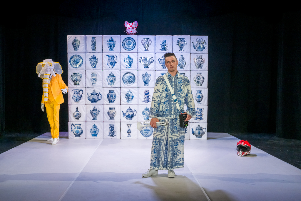 Schauspieler in blau -weißem Anzug steht auf einer Bühne. Im Hintergrund ein weiterer Schauspieler mit Elefantenmaske. Beide stehen vor einer Wand aus weiß-blauen Würfeln. Über die Wand ragt eine Mäusemaske hinaus.