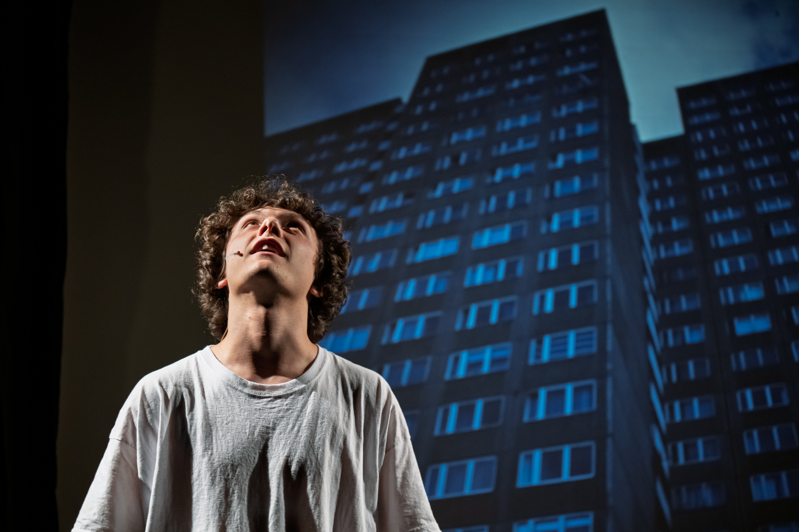 Junger Mann mit lockigem Haar und weißem T-Shirt vor einem Hochhaus mit vielen Fenstern bei Nacht.