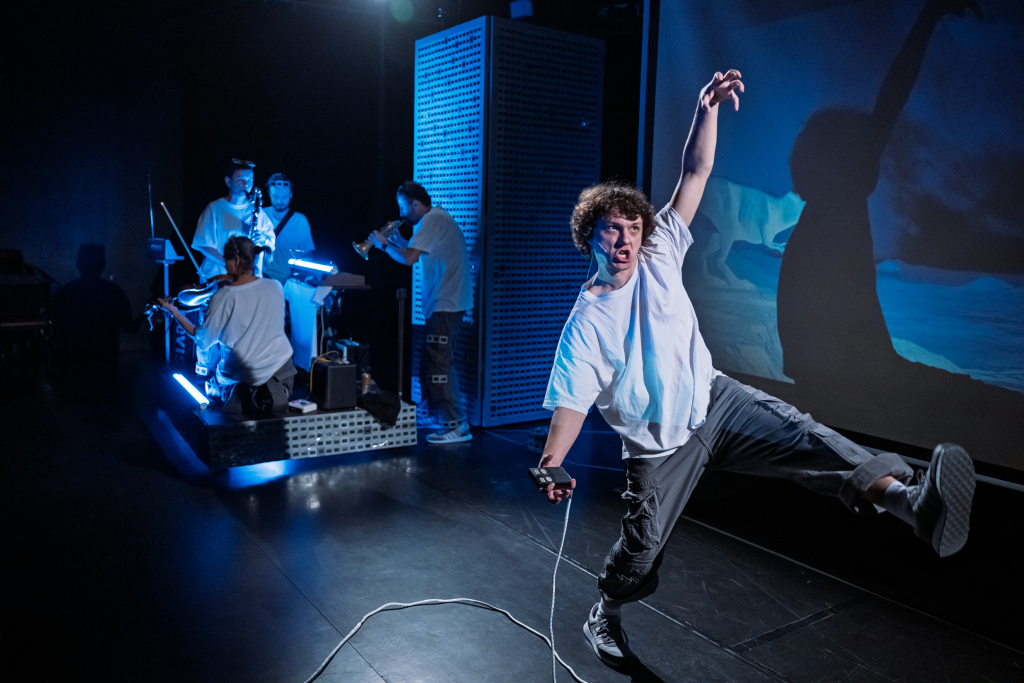 Junge in weißem T-Shirt und dunkler Hose tanzt mit einem Kabel in der Hand vor einer Wand mit Projektion, im Hintergrund sitzen und stehen mehrere Personen an einem beleuchteten Tisch.