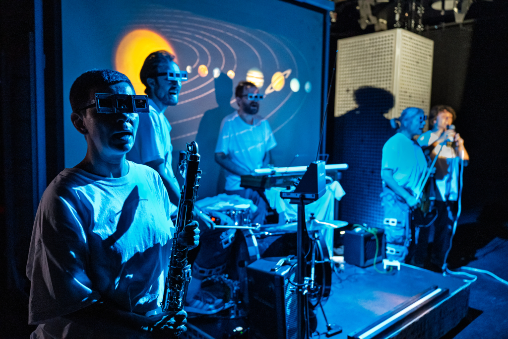 Mehrere Musiker*innen auf einer Bühne mit verschiedenen Instrumenten, im Hintergrund eine Projektion des Sonnensystems.