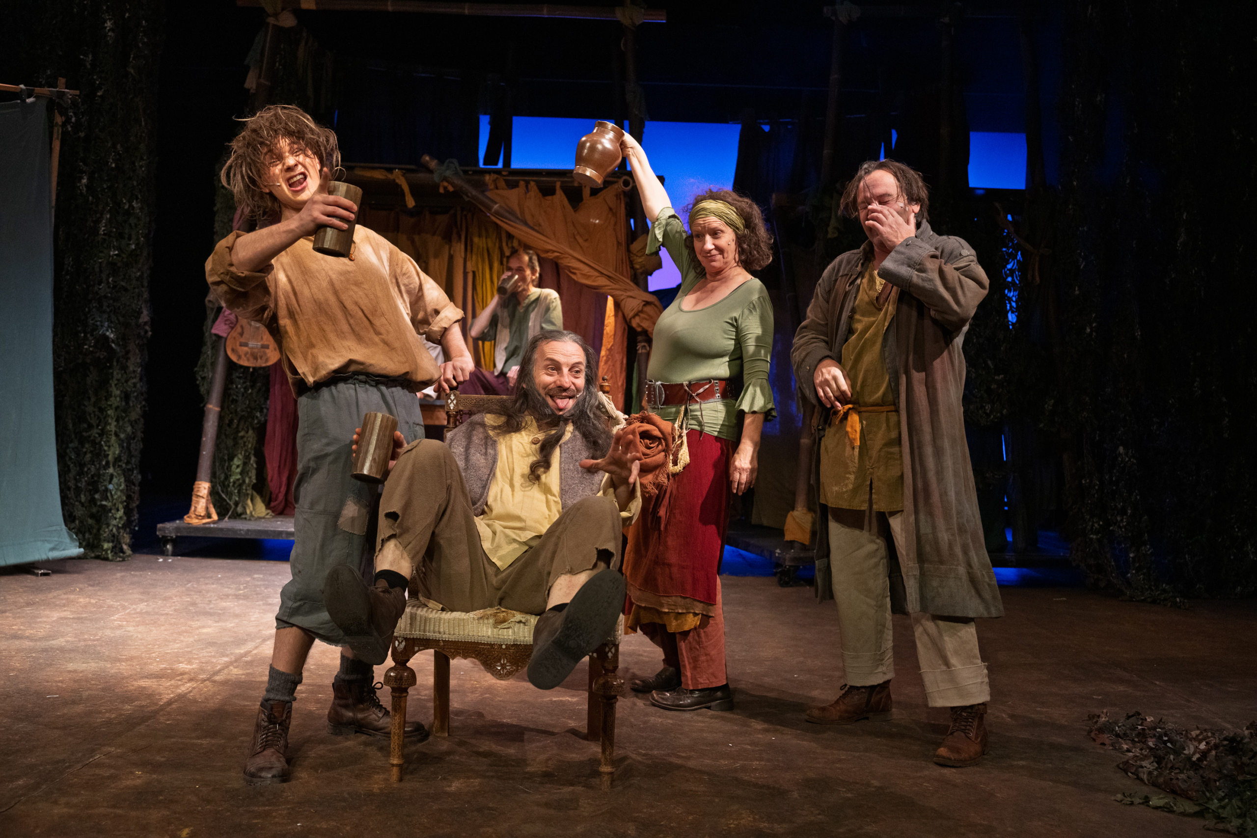 Vier Personen in historischer Kleidung auf einer Bühne, eine sitzt auf einem Stuhl, die anderen stehen, eine hält einen Becher hoch.