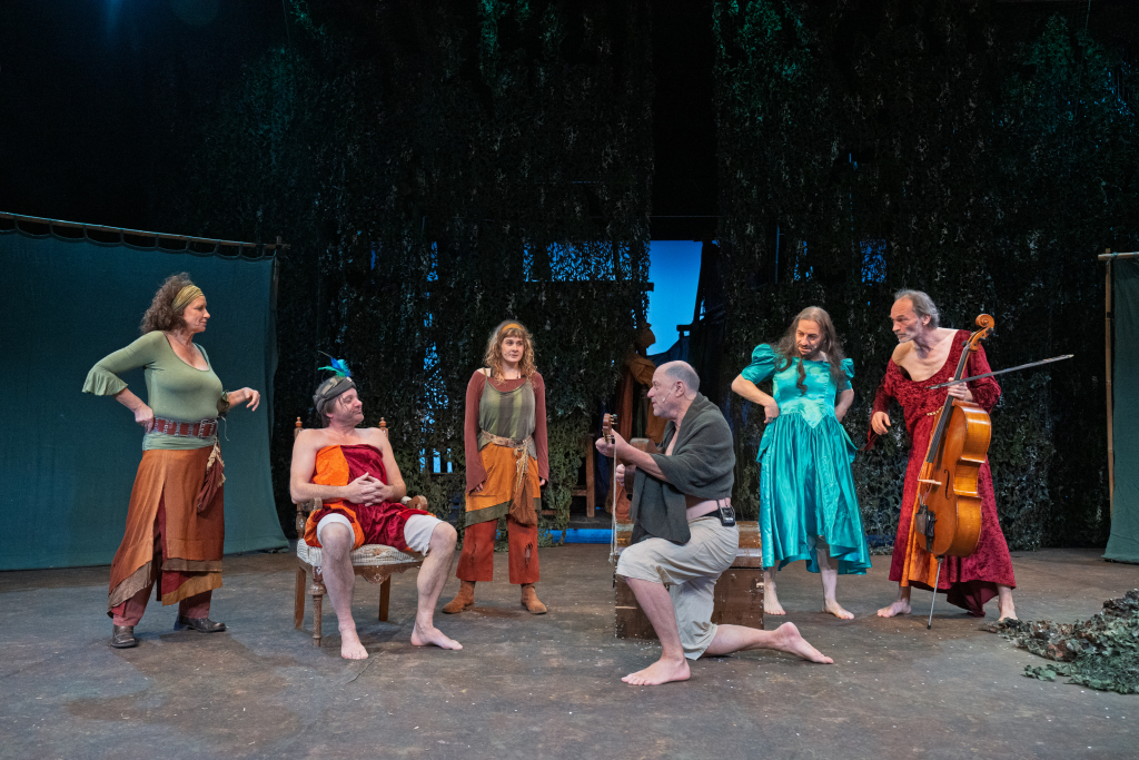 Sechs Personen in bunter, mittelalterlich anmutender Kleidung auf einer Bühne mit Waldkulisse, eine spielt Cello, eine andere Gitarre, eine sitzt auf einem Stuhl.