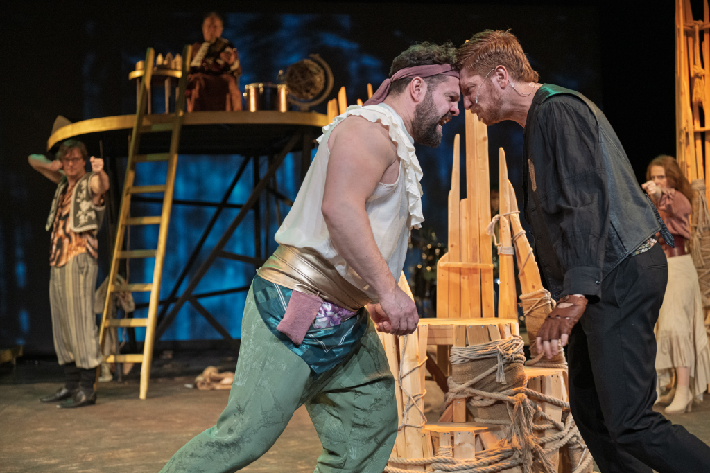 Zwei Männer in historischen Kostümen stehen sich mit den Köpfen nahe vor einem hölzernen Gerüst gegenüber, im Hintergrund weitere Schauspieler auf einer Bühne mit Leiter und Trommel.