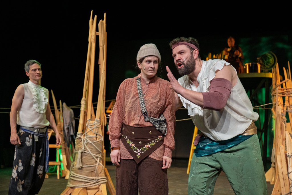 Drei Personen in historisch anmutender Kleidung auf einer Bühne, eine Person gestikuliert mit erhobener Hand, im Hintergrund Bühnenrequisiten aus Holz und Seilen.