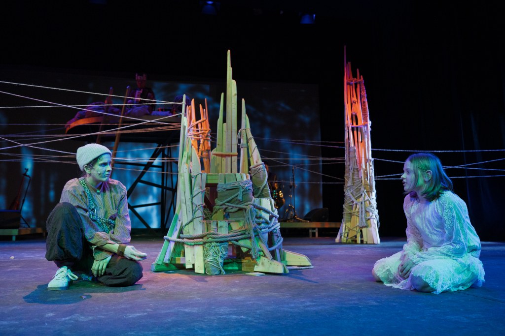 Zwei Personen in bunter Kleidung sitzen auf dem Boden und blicken auf eine aus Holz und Seilen gebaute Skulptur in der Mitte einer Bühne mit dunklem Hintergrund.