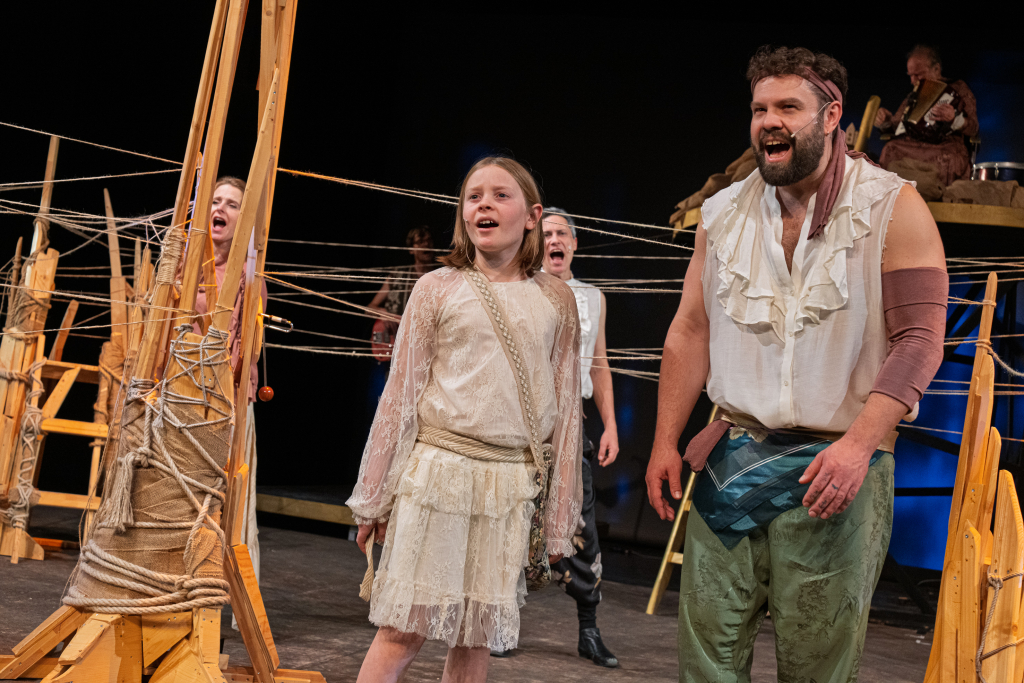 Mehrere Personen in historischen Kostümen stehen auf einer Bühne mit hölzernen Konstruktionen und Seilen im Hintergrund
