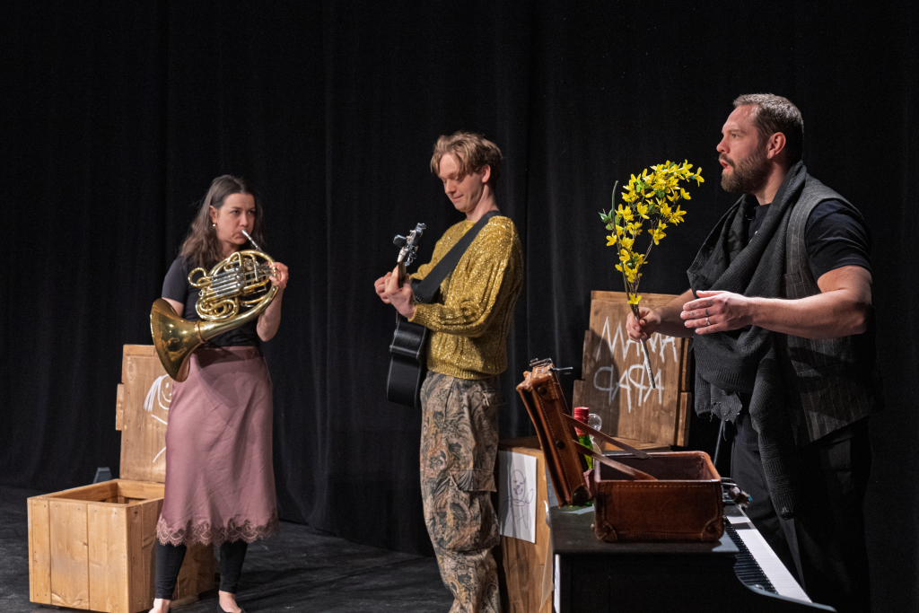 Eine Frau spielt das Horn, ein Mann spielt die Gitarre und ein weiterer Mann hält gelbe Blumen in der Hand.