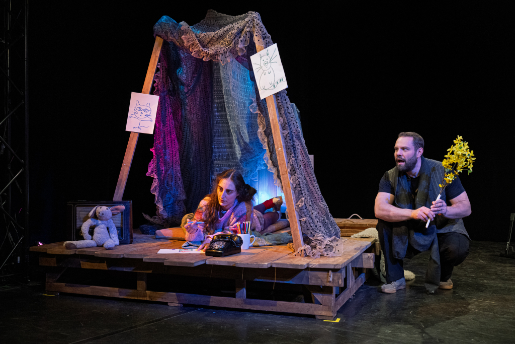 Eine junge Schauspielerin liegt auf dem Bauch in einer aus Decke selsbt gebauten Höhle. Auf der rechten Seite sitzt ein Schauspieler und hält gelbe Blumen in der Hand.