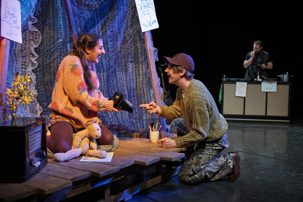 Eine junge Schauspielerin und ein junger Schauspieler grinsen sich an. Die Schauspielerin zeigt dem Schauspieler ein altes Drehscheibentelefon.