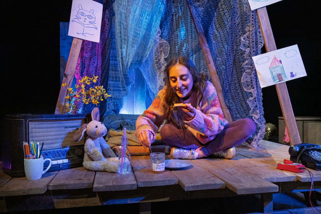 Eine junge Schauspielerin sitzt in einer aus Decken selbstgebauten Höhle. Sie hält eine Waffel mit Marmelade in der Hand. Auf der linken Seite sitzt ihr Kuscheltier.