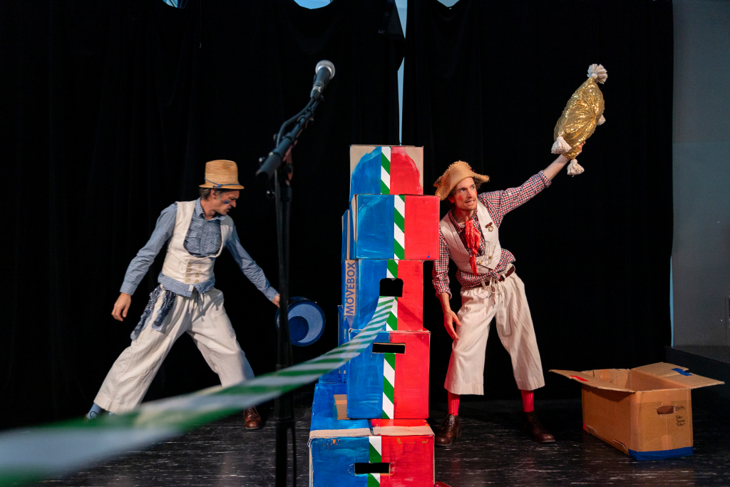Zwei Personen in bunter Kleidung und Hüten auf einer Bühne mit einem Mikrofon und einem Stapel bunter Kartons, eine Person hält ein großes, goldfarbenes Objekt über dem Kopf.