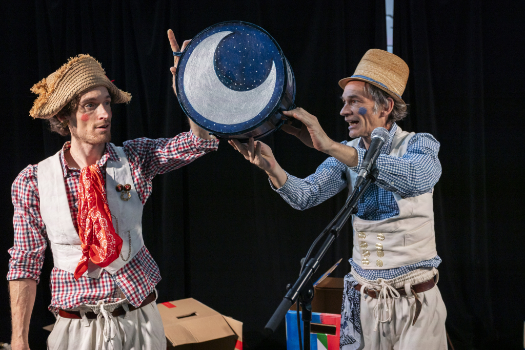 Zwei Personen in karierten Hemden, Westen und Strohhüten halten gemeinsam eine blaue Trommel mit einem weißen Halbmond darauf.