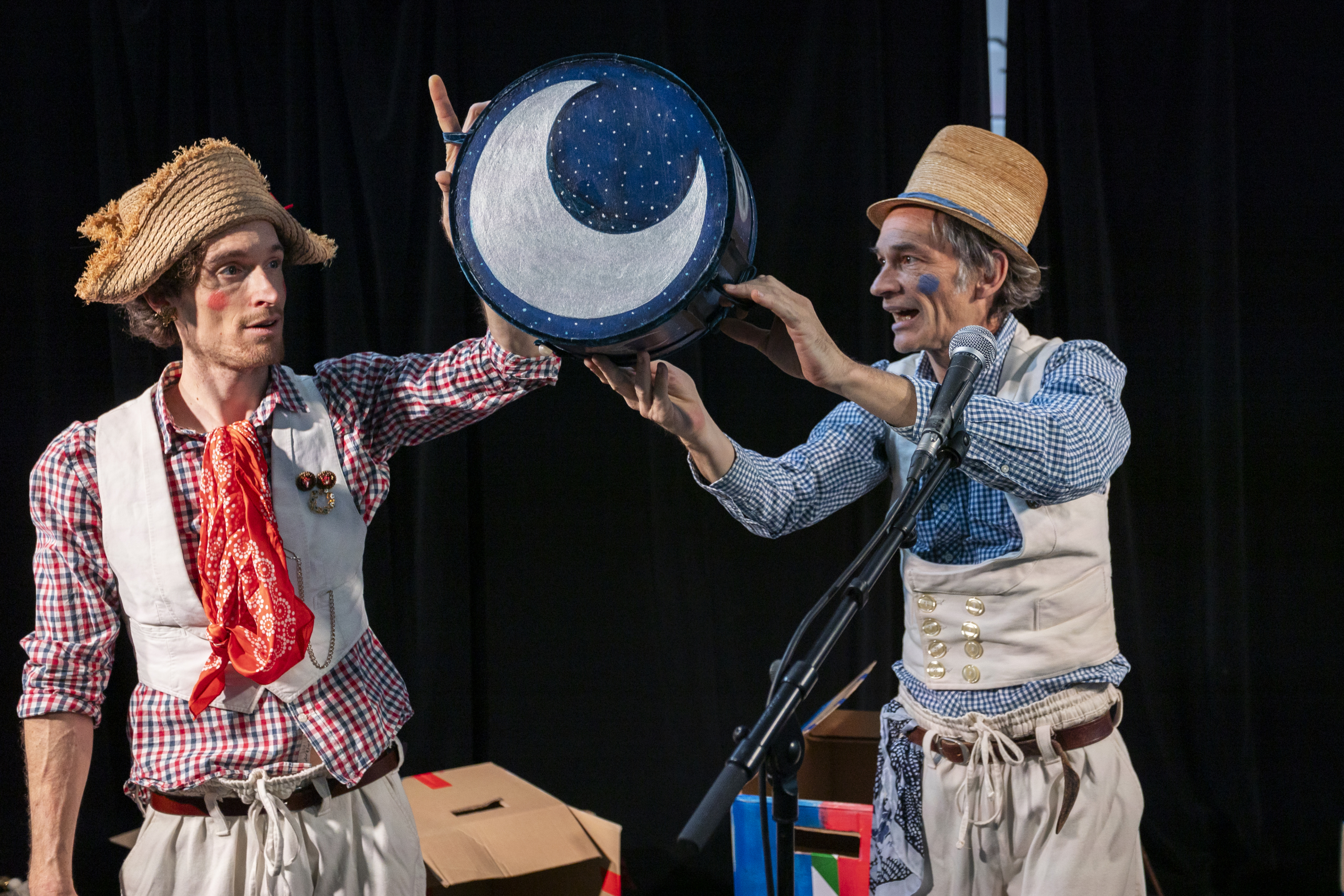 Zwei Personen in karierten Hemden, Westen und Strohhüten halten gemeinsam eine blaue Trommel mit einem weißen Halbmond darauf.