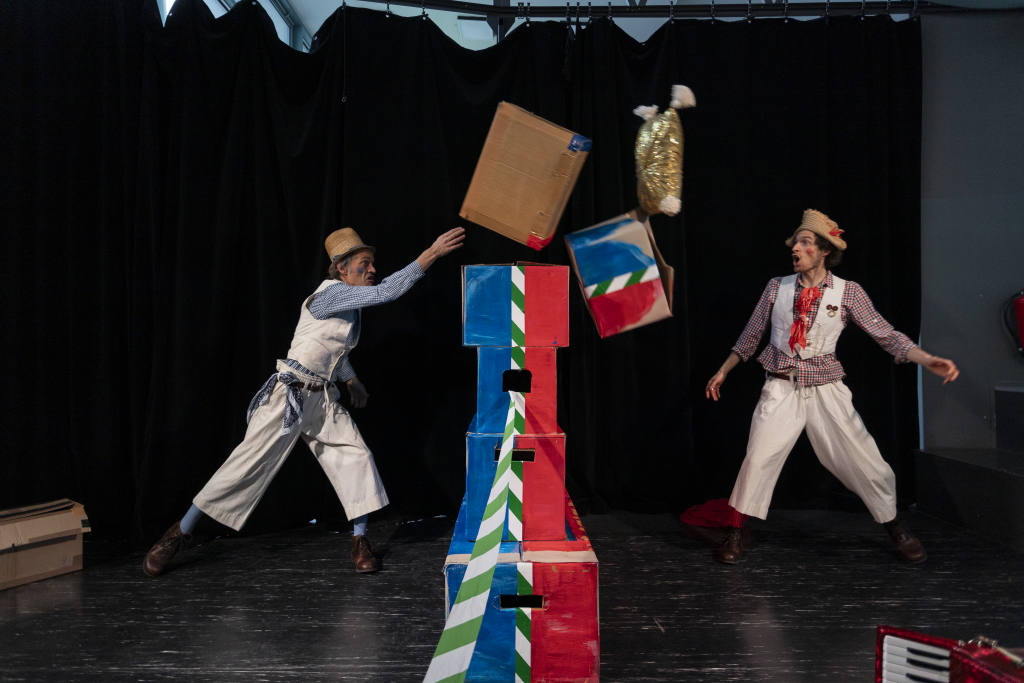 Zwei Personen in weißer Kleidung werfen Gegenstände auf einen Turm aus bunten Kisten auf einer Bühne mit schwarzem Hintergrund.