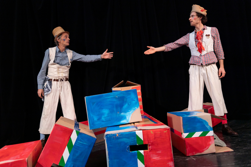 Zwei Personen in bunter, historisch anmutender Kleidung stehen auf einer Bühne mit bemalten Holzkisten.