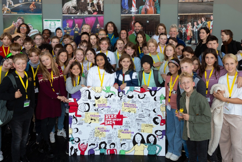 Gruppe von Menschen steht auf einer großen Treppe hinter einem großen, bunten Plakat mit Zeichnungen und Texten.