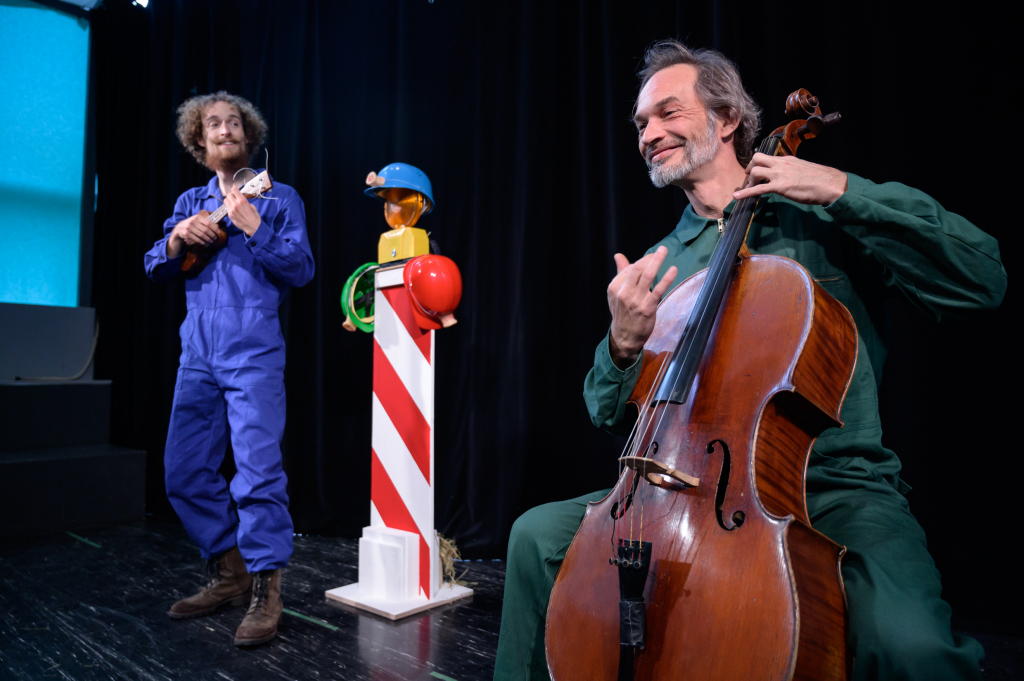 Zwei Musiker auf einer Bühne: einer mit Cello, der andere mit Ukulele, zwischen ihnen ein Baustellensignalschild.