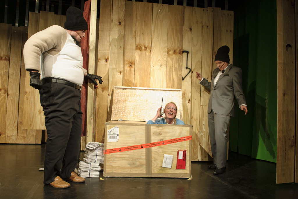 Szenenbild aus einem Theaterstück mit drei Schauspieler*innen, eine schaut aus einer Kiste raus, Freude und Humor sichtbar.