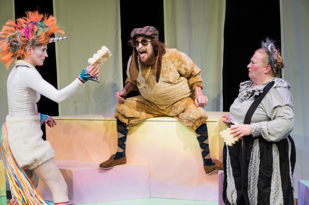Szene aus dem Theaterstück mit drei Schauspielern in bunten Kostümen auf einer farbenfrohen Bühne.