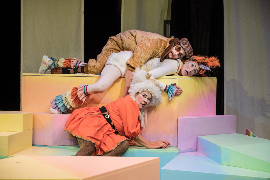 Theateraufführung mit drei Schauspielern in bunten Kostümen auf einer geometrischen Bühne.