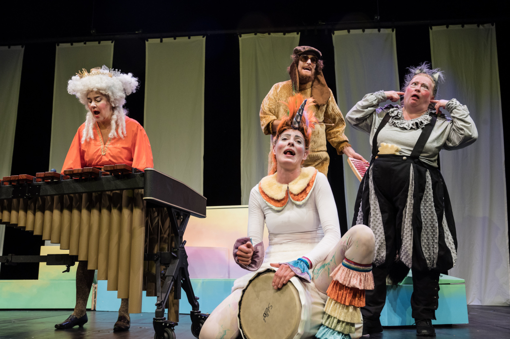 Szenenbild aus einer Theateraufführung mit vier dargestellten Charakteren in bunten Kostümen und Instrumenten.