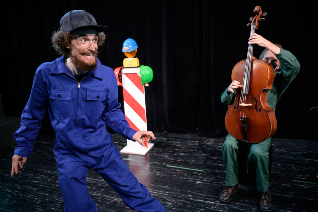 Schauspieler in blauer Arbeitskleidung während einer Aufführung, mit einem Cellisten im Hintergrund.