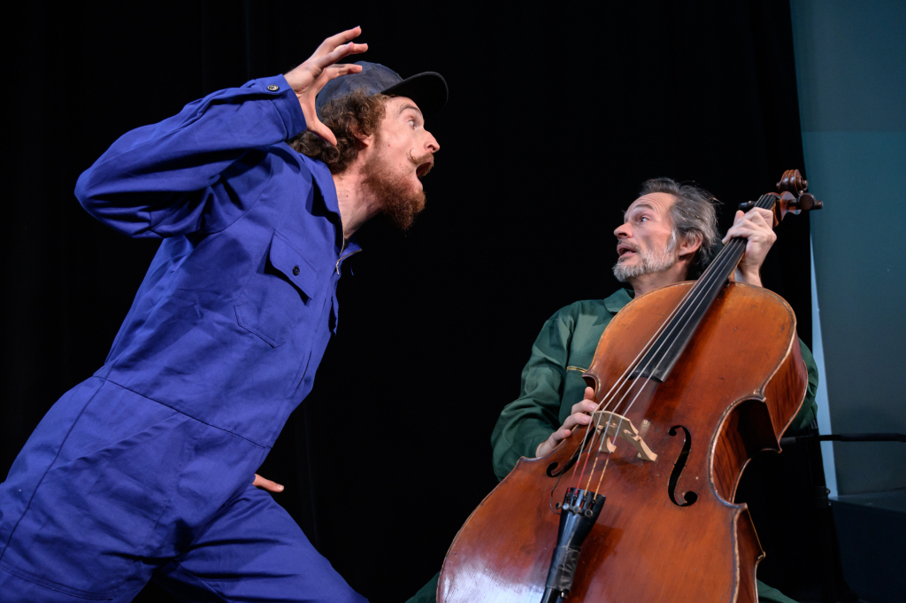 Szenenbild aus einem Theaterstück mit zwei Schauspielern, einer erschreckt den anderen, der andere spielt Cello und schrickt zurück.