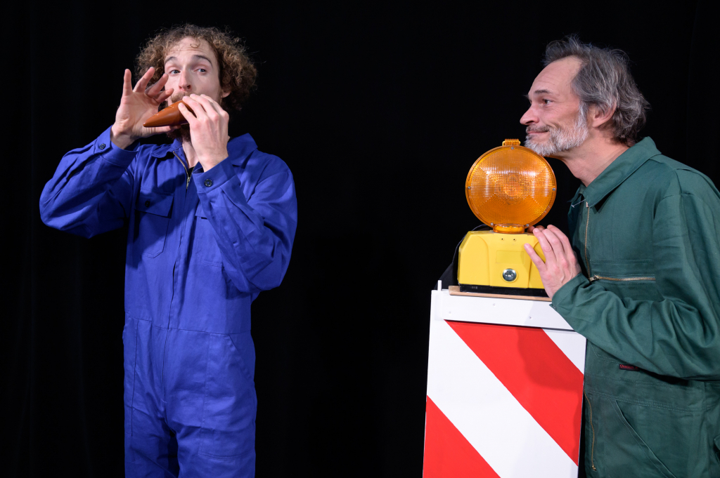 Zwei Schauspieler in Arbeitskleidung, einer spielt eine Mundharmonika, der andere lächelt neben einer Warnleuchte.