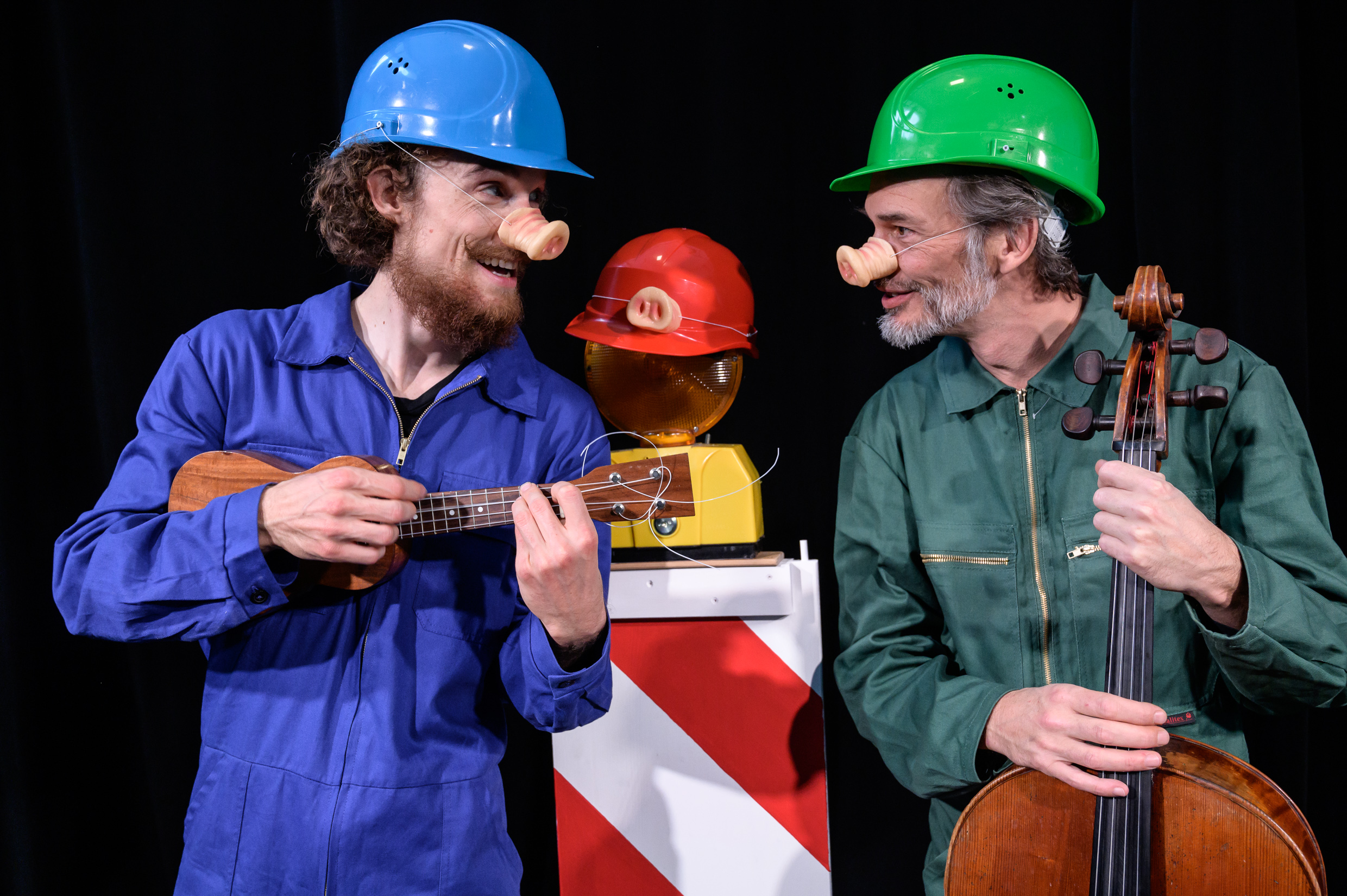 Zwei Musiker mit Schweinenasen: Einer spielt Ukulele, der andere Cello, mit Baustellensignalschild im Hintergrund.