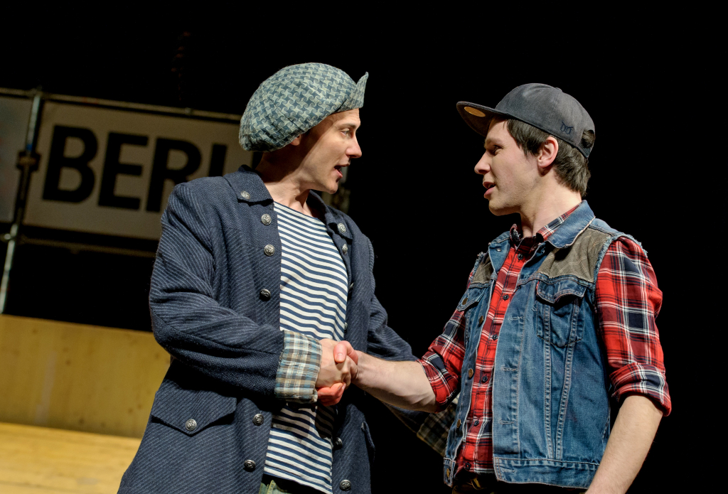 Zwei Schauspieler geben sich die Hand. Im Hintergrund ein Schild mit der Aufschrift "BERLIN"