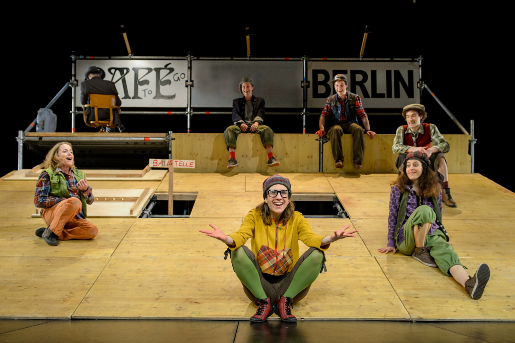 Szenenbild mit sechs Personen auf einer Holzbühne, fröhliche Darstellung auf der Bühne.