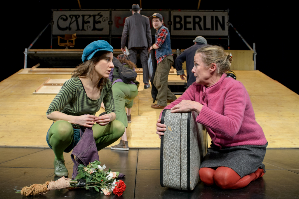 Zwei Schauspielerinnen im Vordergrund und einer Gruppe von Schauspielern im Hintergrund.