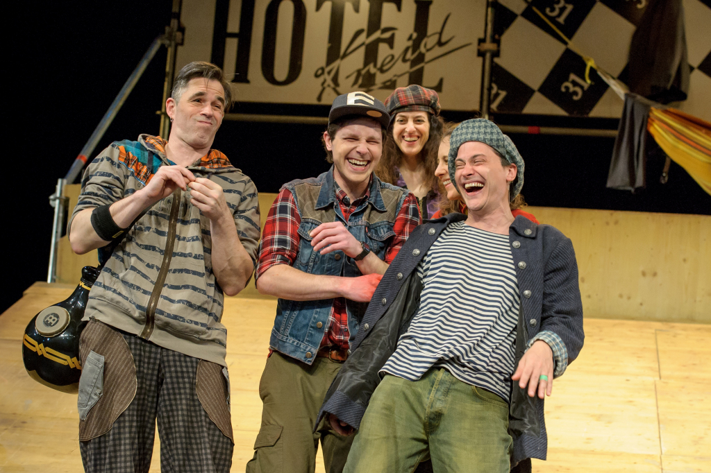 Szenenbild mit vier lachenden Schauspieler*innen auf der Bühne. Im Hintergrund ein Hotelschild.