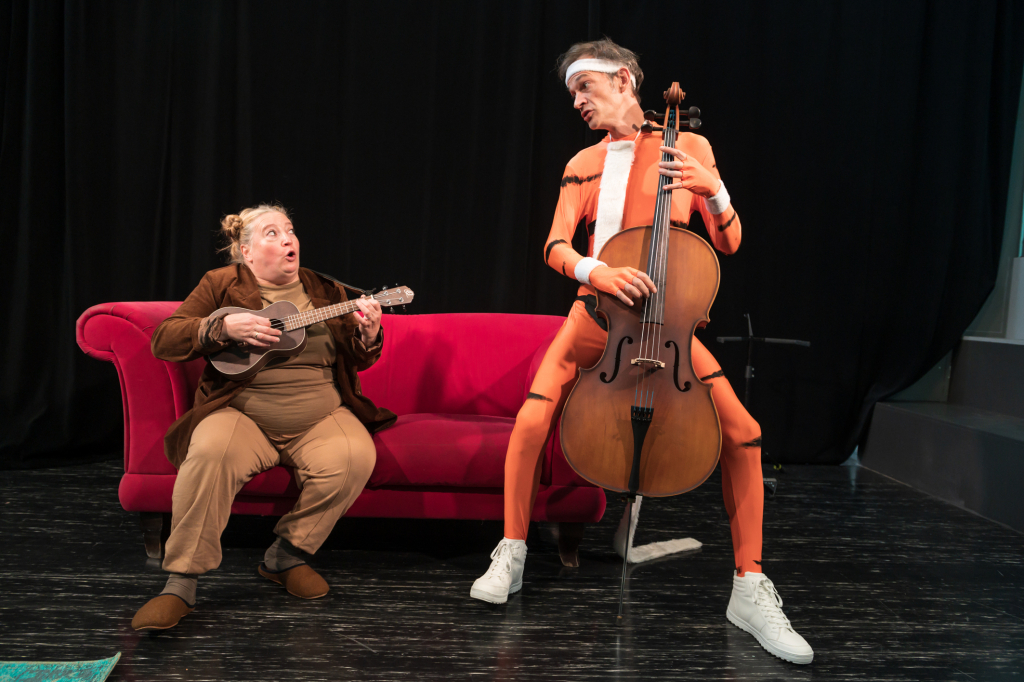Zwei Personen auf einer Bühne: eine sitzt auf einem roten Sofa und spielt Ukulele, die andere steht und spielt Cello, trägt orangefarbenen Ganzkörperanzug mit schwarzen Streifen.