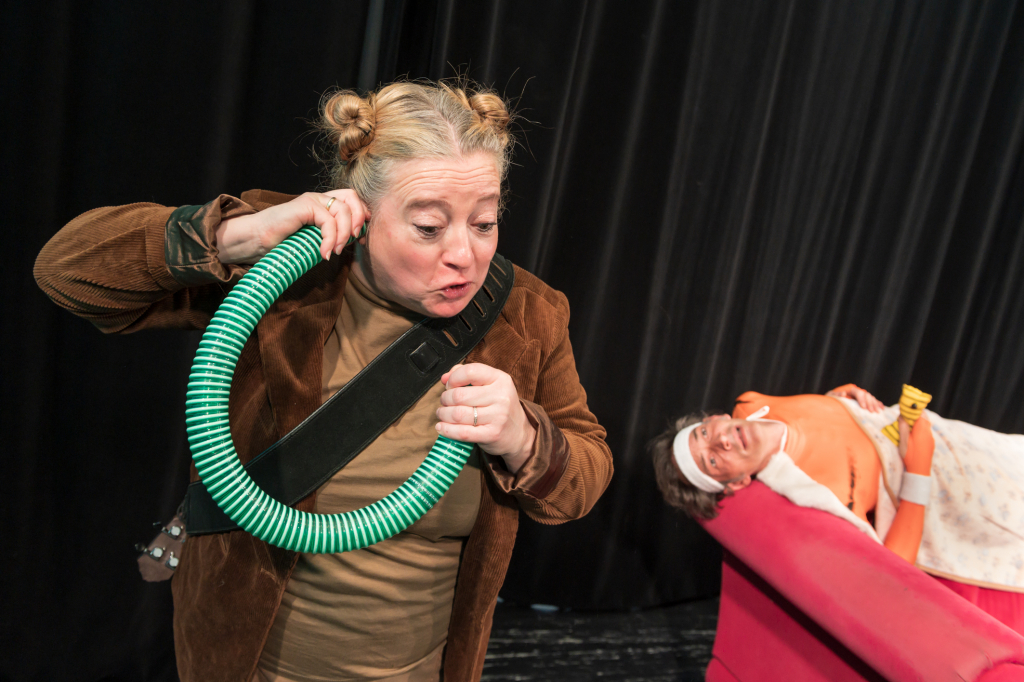 Person mit brauner Jacke hält grünen, flexiblen Schlauch, im Hintergrund liegt eine weitere Person auf einem roten Sofa mit Decke und Kissen.